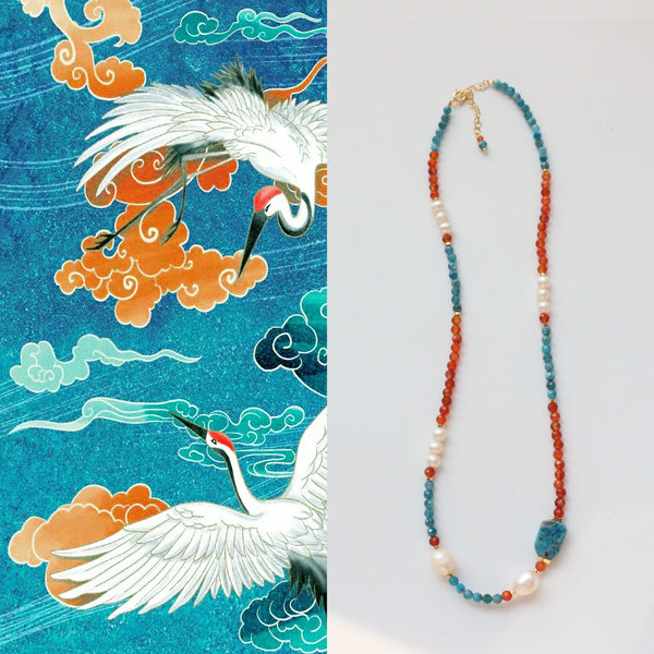 Image of the Azure Flame Necklace featuring a vibrant blend of blue apatite, red-orange agate beads, and interspersed with elegant white freshwater pearls, set against a textured background illustrating a majestic white crane amidst clouds