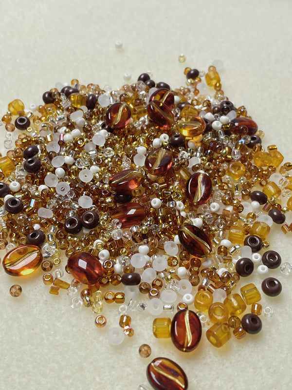 An assortment of warm amber-toned acrylic and glass beads in various shapes and sizes, including translucent and matte finishes, displayed in a scoop and a decorative white bowl. Ideal for DIY jewelry making, the beads showcase rich earthy colors, adding a touch of warmth and charm to handmade bracelets and necklaces.